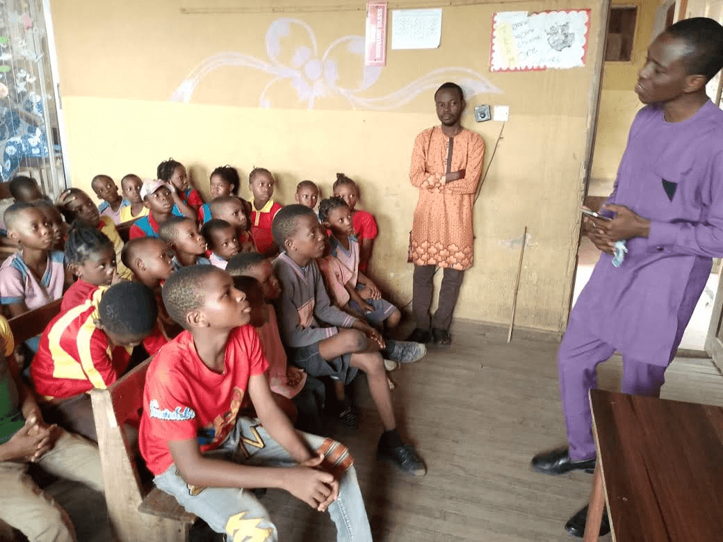 Two men teaching a classroom of seated children, who are looking up at them.