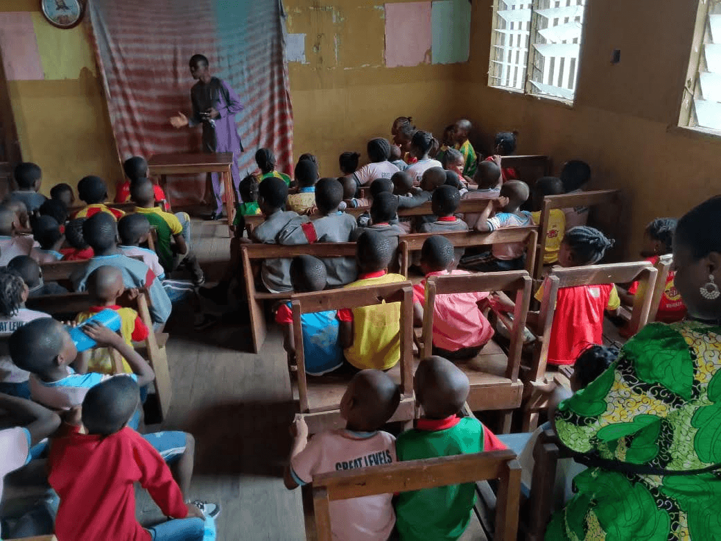 A wide view of the classroom, rows of children at wooden desks facing a man teaching at the front.