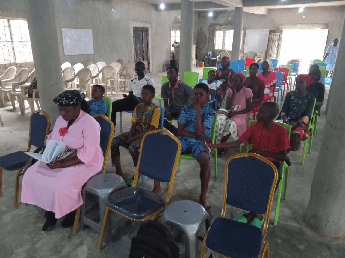 Children and adults seated on plastic chairs in an unfinished hall, listening to the session.