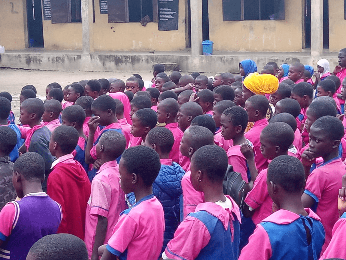 A crowd of primary school pupils in pink and blue uniforms standing at assembly in a school yard.