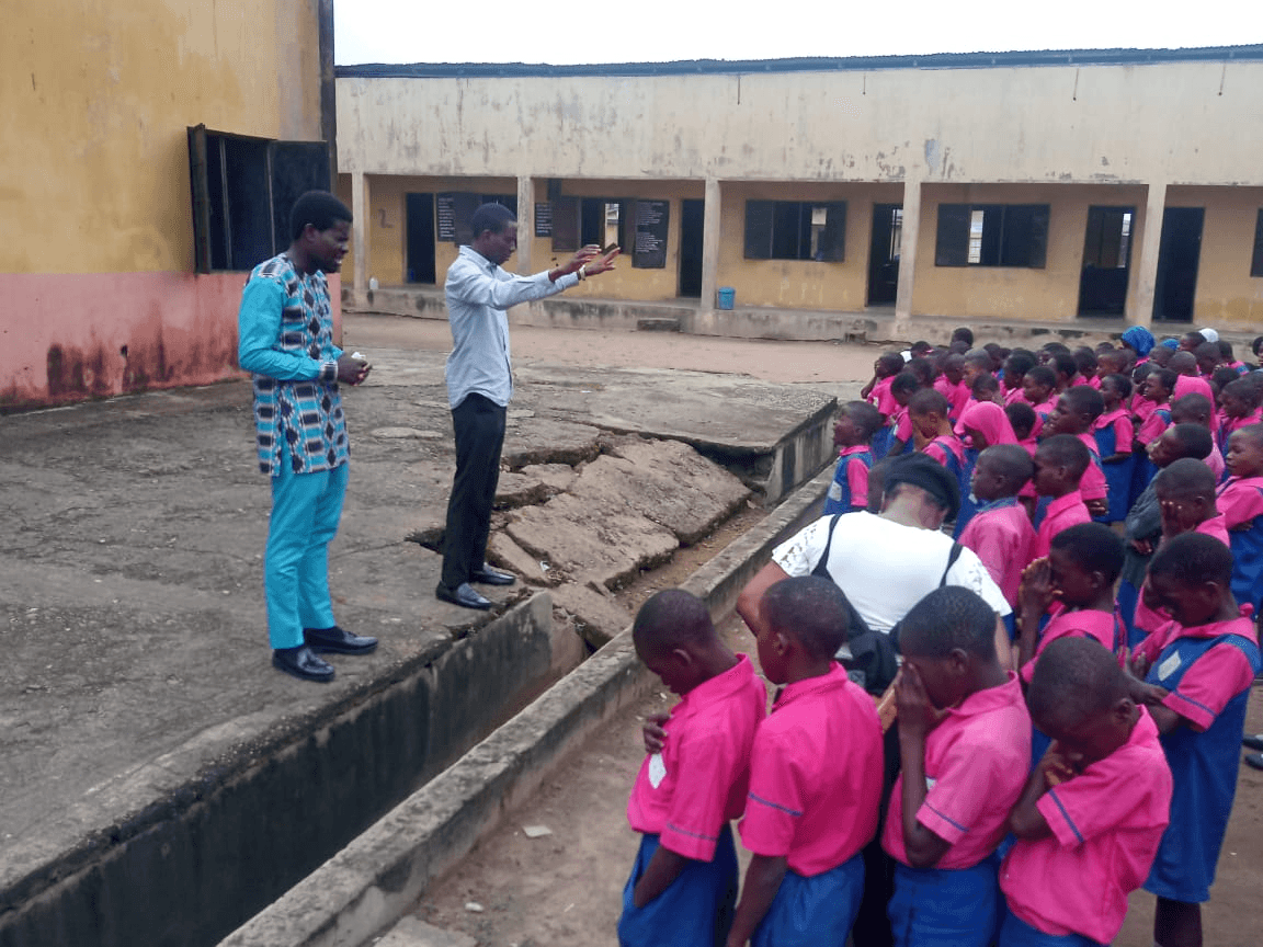 Two men speaking to rows of pupils gathered along a low wall in the school yard.