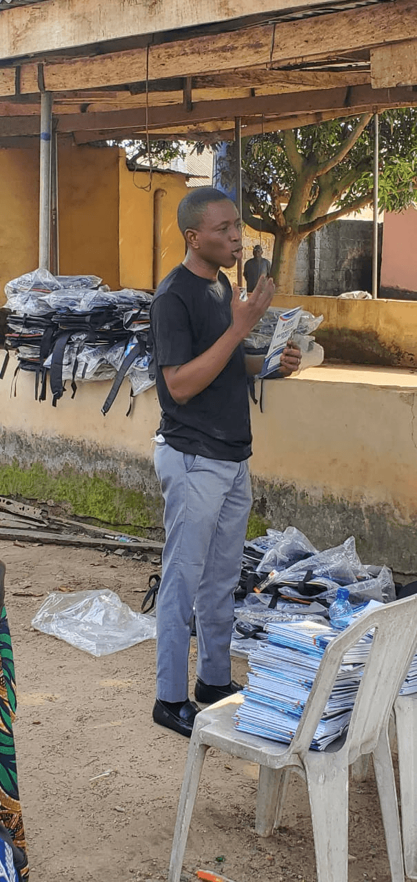 A man speaking while holding a booklet, with stacks of exercise books and bagged school bags behind him.