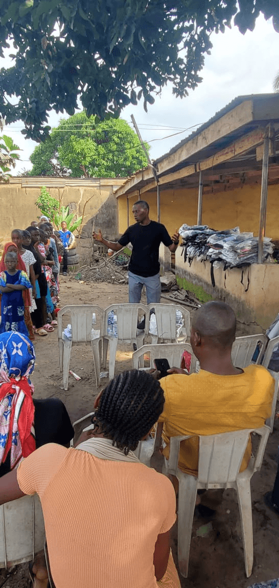 A man addressing parents and children seated on plastic chairs in a yard.